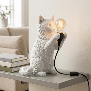 Op de afbeelding: Een witte tafellamp in de vorm van een kat met een textuur van lang haar, staand op de achterpoten. De kat houdt een zwarte fitting vast met een gloeiende, heldere Edison-gloeilamp. Een zwart stroomsnoer met een schakelaar steekt naar buiten.