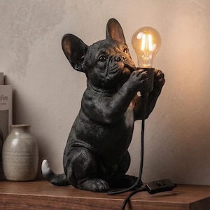 Peut inclure: Une lampe figurine de bouledogue français noir tenant une ampoule de style Edison allumée dans ses pattes avant. Le chien a des oreilles droites et une queue à bout blanc. Un cordon noir avec interrupteur part de la base de l'ampoule.