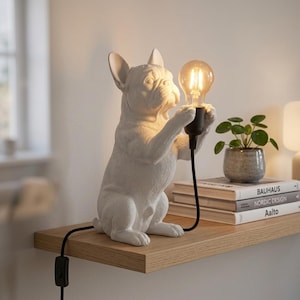 Op de afbeelding: Een witte tafellamp in de vorm van een Franse bulldog die op zijn achterpoten staat en een brandende Edison-gloeilamp in zijn voorpoten houdt. De lamp is voorzien van een zwart stroomsnoer met een ingebouwde schakelaar voor bediening.