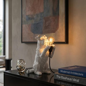 Op de afbeelding: Een wit, getextureerd hondenbeeldje dat op zijn achterpoten staat en een zwarte fitting met een gloeiende, warmgekleurde Edison-lamp vasthoudt. De hond heeft een ruige vacht en staat naast een kleine glazen vaas en twee opgestapelde boeken.