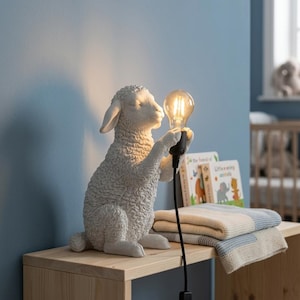 Op de afbeelding: Een witte, getextureerde tafellamp in de vorm van een schaap die een heldere Edison-stijl gloeilamp in de voorpoten houdt. De lamp heeft een zwart netsnoer met een schakelaar en geeft warm licht via de gloeilamp die het schaap vasthoudt.