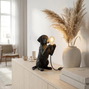 May include: A black dog-shaped table lamp featuring a seated puppy holding a single exposed Edison-style light bulb in its front paws. The lamp includes a black power cord with an inline switch, providing a focused light source for interior spaces.