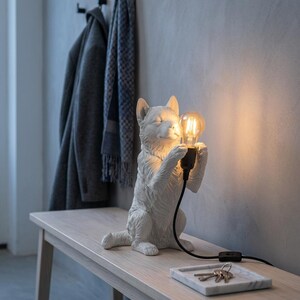 Op de afbeelding: Een witte, sculpturale hondfiguurlamp die rechtop zit en een gloeiende, heldere Edison-stijl gloeilamp in de voorpoten houdt. De lamp heeft een zwart netsnoer met een schakelaar, waardoor het gezicht van de hond warm wordt verlicht.