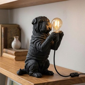 Op de afbeelding: Een matzwarte tafellamp in de vorm van een zittende Shar-Pei hond met een gerimpelde huid. Het hondenfiguur houdt een heldere glazen Edison-gloeilamp tussen de voorpoten vast. Een zwart netsnoer met een schakelaar loopt vanaf de voet van de lamp.