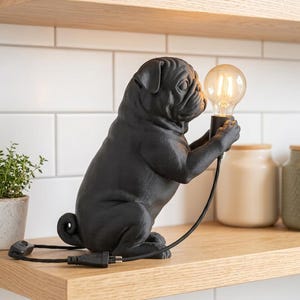 May include: A matte black pug-shaped table lamp featuring a seated dog figure holding a clear glass Edison-style light bulb in its front paws. The lamp includes a black power cord with an inline switch and a standard two-prong electrical plug.
