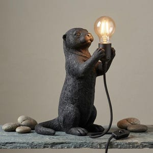 Può includere: Una lampada da tavolo a forma di lontra nera, in posizione eretta, che tiene una lampadina in vetro trasparente tra le zampe anteriori. La lampada presenta una finitura scura testurizzata, un cavo nero con interruttore e una lampadina.