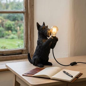 Op de afbeelding: Een zwarte Scottish Terrier hondfiguur lamp die een gloeiende Edison-stijl gloeilamp in zijn voorpoten houdt. De lamp heeft een zwart snoer met een schakelaar en staat naast een open notitieboek met een zwarte pen op een houten bureau.