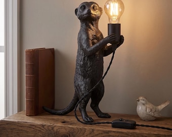 Peut inclure: Une lampe de table noire en forme de suricate debout, tenant une douille avec une ampoule transparente de style Edison. La lampe présente une surface texturée imitant la fourrure, une longue queue et un cordon d'alimentation noir avec interrupteur.