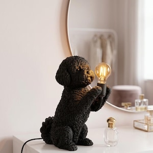 Op de afbeelding: Een zwarte tafellamp in de vorm van een zittende poedel met een krullende vachtstructuur. Het hondenfiguur houdt tussen de voorpoten een zwarte fitting vast met een gloeiende, ronde, heldere glazen lamp in Edison-stijl. Zwart snoer.