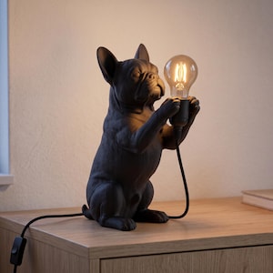 Op de afbeelding: Een matzwarte tafellamp in de vorm van een Franse Bulldog die op zijn achterpoten staat. De hond houdt een brandende lamp in Edison-stijl vast met zijn voorpoten. Een zwart netsnoer met een schakelaar loopt vanaf de voet van de lamp.