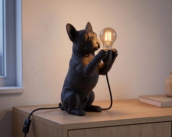 Op de afbeelding: Een matzwarte tafellamp in de vorm van een Franse Bulldog die op zijn achterpoten staat. De hond houdt een brandende lamp in Edison-stijl vast met zijn voorpoten. Een zwart netsnoer met een schakelaar loopt vanaf de voet van de lamp.