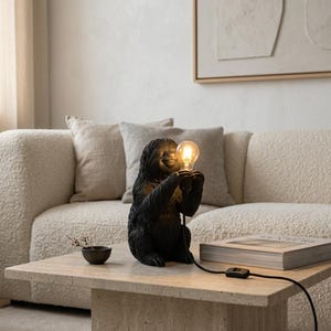 Puede incluir: Una lámpara de mesa con forma de perezoso negro, acabado texturizado y postura sentada. El perezoso sostiene entre sus manos una bombilla de cristal transparente que emite una luz cálida. Incluye un cable de alimentación negro con interruptor.