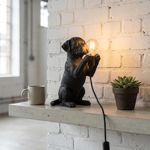 Op de afbeelding: Een zwarte tafellamp in de vorm van een hond met een sculptuur van een zittende puppy die een heldere glazen gloeilamp met een zichtbare gloeidraad vasthoudt. De lamp heeft een zwart netsnoer met een schakelaar aan de basis.