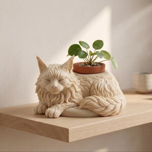 Op de afbeelding: Een crèmekleurige, sculpturale plantenbak in de vorm van een rustende langharige kat met gesloten ogen. Het kattenfiguur heeft een gedetailleerde vachttextuur en een ronde opening op de rug met een kleine terracotta pot met een Pilea-plant.