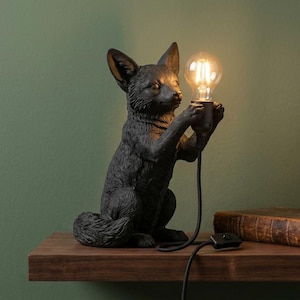 May include: A matte black fox-shaped table lamp featuring a textured fur finish. The fox is depicted in a seated position, holding a clear glass LED light bulb between its front paws. A black power cord with an inline switch extends from the base.