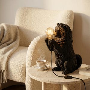 Op de afbeelding: Een zwarte tafellamp in de vorm van een hond met een getextureerde vacht in zittende houding. De hond houdt een heldere, gloeiende Edison-stijl lamp vast tussen de voorpoten. Een zwart snoer met een schakelaar loopt vanaf de lamp.