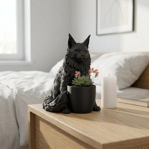 Puede incluir: Una maceta con forma de gato en color negro mate que presenta una figura felina detallada y texturizada con orejas puntiagudas y pelaje largo. El gato está sentado erguido y sostiene una maceta cilíndrica negra lisa con una suculenta verde.