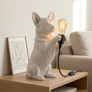 Op de afbeelding: Een witte tafellamp met een matte afwerking in de vorm van een zittende Corgi-hond. Het hondenfiguur houdt in zijn voorpoten een zwarte fitting met een gloeiende Edison-stijl lamp vast. Een zwart netsnoer met schakelaar loopt vanaf de voet.