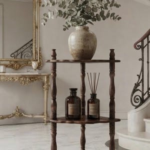 May include: A three-tiered dark brown wooden corner stand with scalloped edges on each shelf. The top shelf holds a speckled beige ceramic vase with eucalyptus branches, while the middle shelf displays two amber glass bottles with labels.