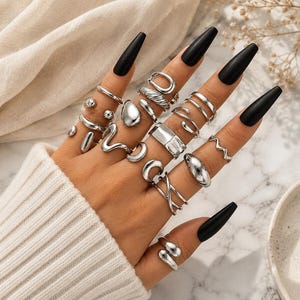 May include: A collection of multiple silver-toned metal rings worn on fingers. The rings feature various designs including smooth bands, twisted textures, open shapes, wavy lines, and polished surfaces. The rings are worn across all five fingers.