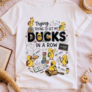 May include: A white t-shirt featuring a colorful graphic of yellow ducklings in a classroom setting. The text reads "Trying to get my ducks in a row" with "Mrs. Jones" written near the ducks. The design includes books, alphabet blocks, a chalkboard, and a bell.