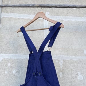 May include: A pair of deep blue denim overalls hanging on a wooden hanger. The garment features wide shoulder straps that cross at the back, metal hardware adjusters, and a large front bib pocket. The fabric has a visible texture and deep blue color.