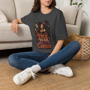 May include: A charcoal grey t-shirt featuring a graphic illustration of a witch wearing a hat and holding a steaming mug. The text reads Hex Yeah, I Need Coffee in orange and purple stylised lettering, accompanied by blue jeans and white trainers.