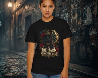 May include: A black t-shirt featuring a graphic of a black cat wearing a top hat, surrounded by red roses and a crescent moon. Below the cat, the text reads "THE DARK KNOWS MY NAME" in a decorative, gold-colored serif font.