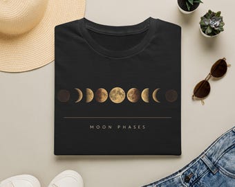 May include: A black t-shirt featuring a horizontal graphic of the moon phases in shades of gold and brown. Below the moon cycle, a thin horizontal line separates the graphic from the text MOON PHASES printed in a clean, sans-serif font.