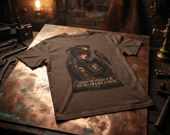 May include: A brown short-sleeved t-shirt featuring a graphic print of a person in a top hat with a raven and a heart, accompanied by the text: I BURIED THE VERSION OF ME THAT ONLY KNEW HOW TO ENDURE. The design is centered on the chest area.