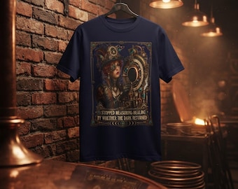 May include: A navy blue t-shirt featuring a graphic print of a person wearing a hat and goggles, surrounded by mechanical gears and clockwork elements. The text below the illustration reads: STOPPED MEASURING HEALING BY WHETHER THE DARK RETURNED.