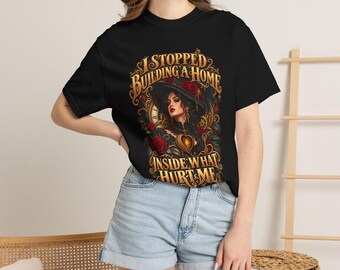 May include: A black short-sleeve t-shirt featuring a graphic print of a person in a wide-brimmed hat surrounded by red roses and gold clockwork elements, with the text I STOPPED BUILDING A HOME INSIDE WHAT HURT ME.