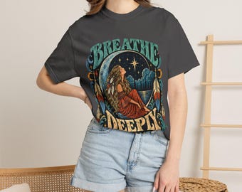 May include: A dark gray short-sleeve t-shirt featuring a graphic print of a person sitting under a crescent moon and stars with the text BREATHE DEEPLY. The design includes feathers, clouds, and a body of water in shades of blue, red, and gold.