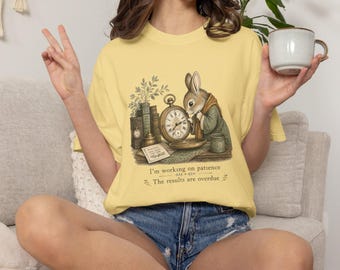 May include: A pale yellow t-shirt featuring a graphic of a rabbit wearing a scarf, standing next to a stack of books, a pocket watch, and a small mug. The text below the illustration reads, I'm working on patience. The results are overdue.