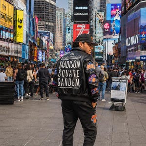 May include: A black leather bomber jacket with white text reading Madison Square Garden, the worlds most famous arena, and colorful patches on the sleeve. The jacket is paired with black joggers and white sneakers with orange accents and a black baseball cap.