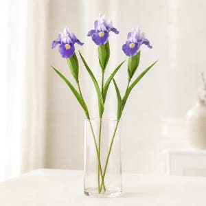 May include: Three artificial iris flowers with purple and light blue petals, yellow centers, and green stems, arranged in a clear cylindrical glass vase. The flowers feature detailed, layered petals and long, slender green leaves on each stem.