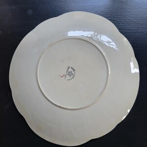 May include: The underside of a cream-colored ceramic plate with a scalloped edge. In the center, a black circular maker's mark is visible, featuring the text 'MADE IN ENGLAND' and a small emblem, along with a small red handwritten mark.