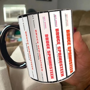 May include: A white ceramic mug featuring a graphic print of five cassette tape spines. The spines display the text Bruce Springsteen and album titles including Human Touch, Lucky Town, Tunnel of Love, Born in the U.S.A., and Nebraska in red lettering.