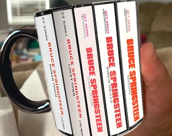Puede incluir: Una taza de cerámica blanca con un estampado gráfico de cinco lomos de casete. Los lomos muestran el texto Bruce Springsteen y los títulos de álbumes Human Touch, Lucky Town, Tunnel of Love, Born in the U.S.A. y Nebraska en letras rojas.