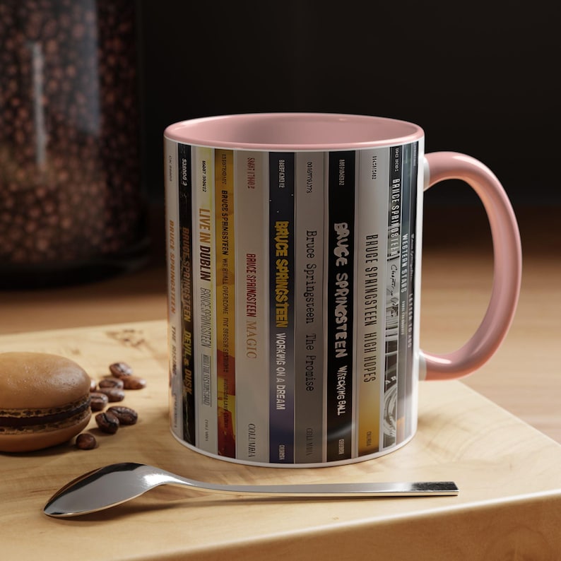 May include: A ceramic mug with a light pink interior and handle, featuring a wrap-around graphic of various Bruce Springsteen album spines. The design displays titles like Live in Dublin, Magic, Working on a Dream, The Promise, and High Hopes.