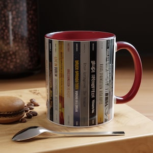 May include: A ceramic mug with a deep red interior and handle, featuring a wrap-around graphic of various Bruce Springsteen album spines. The design displays titles like Live in Dublin, Magic, The Promise, Wrecking Ball, High Hopes, and Western Stars.
