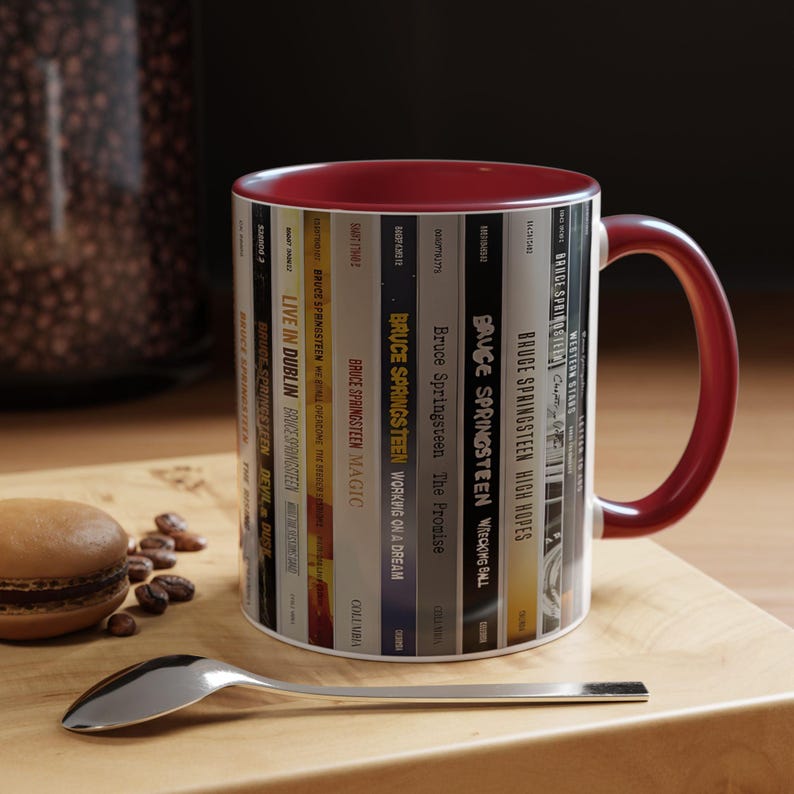 May include: A ceramic mug with a deep red interior and handle, featuring a wrap-around graphic of various Bruce Springsteen album spines. The design displays titles like Live in Dublin, Magic, The Promise, Wrecking Ball, High Hopes, and Western Stars.