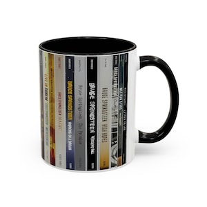 May include: A white ceramic mug with a black interior and handle, featuring a wrap-around graphic of various Bruce Springsteen album spines including Live in Dublin, Magic, Working on a Dream, The Promise, Wrecking Ball, High Hopes, and Western Stars.