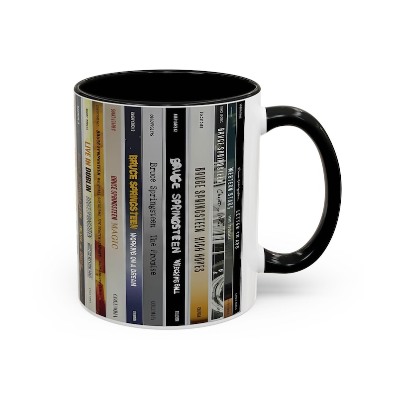 May include: A white ceramic mug with a black interior and handle, featuring a wrap-around graphic of various Bruce Springsteen album spines including Live in Dublin, Magic, Working on a Dream, The Promise, Wrecking Ball, High Hopes, and Western Stars.
