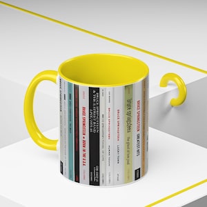 May include: A ceramic mug with a bright yellow interior and handle. The exterior features a vertical graphic design displaying a series of Bruce Springsteen album spines, including titles like Born in the U.S.A., The River, and Born to Run.