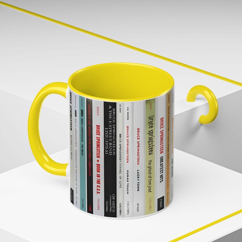 May include: A ceramic mug with a bright yellow interior and handle. The exterior features a vertical graphic design displaying a series of Bruce Springsteen album spines, including titles like Born in the U.S.A., The River, and Born to Run.