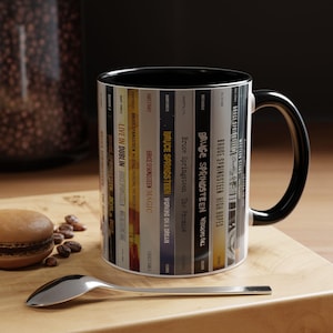 May include: A white ceramic mug with a black interior and handle, featuring a wrap-around graphic of various Bruce Springsteen album spines. The design includes text titles like Live in Dublin, Magic, The Promise, Wrecking Ball, and High Hopes.