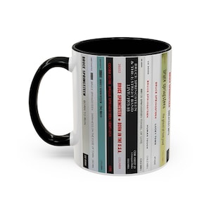 May include: A white ceramic mug with a black handle and interior, featuring a vertical graphic design of multiple Bruce Springsteen album spines. The text includes titles like Born to Run, The River, Nebraska, Born in the U.S.A., and Greatest Hits.