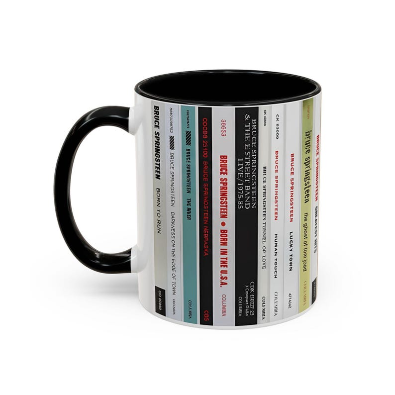 May include: A white ceramic mug with a black handle and interior, featuring a vertical graphic design of multiple Bruce Springsteen album spines. The text includes titles like Born to Run, The River, Nebraska, Born in the U.S.A., and Greatest Hits.