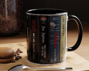 Puede incluir: Una taza de cerámica con interior y asa de color negro, que presenta un diseño gráfico envolvente de varias cintas de casete. Las cintas están etiquetadas con los títulos de los álbumes de Foo Fighters, incluyendo One by One, There Is Nothing Left to Lose y The Colour and the Shape.
